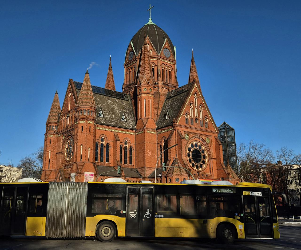 A beautiful red-brick church in Berlin with a yellow city bus in the foreground on a clear sunny day.